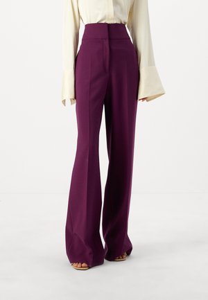 Fioletowe spodnie o wysokim stanie i szerokich nogawkach noszone z kremową, długorękawową bluzką z szerokimi mankietami oraz beżowymi sandałami na odkryte palce.