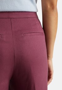 Bordowe spodnie o kroju dopasowanym, z gładką fakturą i kieszeniami bocznymi, widziane z tyłu.