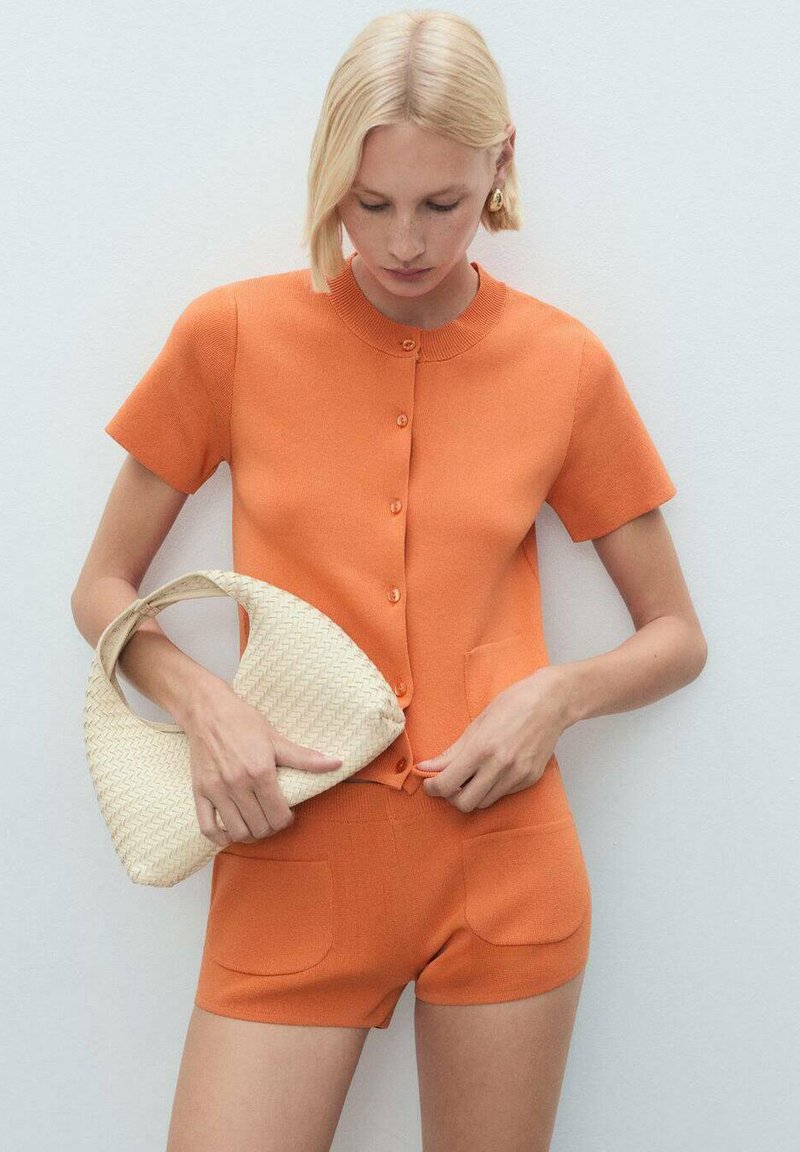 Orange stickad tvådelad outfit bestående av en kortärmad topp med knappar och matchande shorts, tillsammans med en vävd krämfärgad handväska.