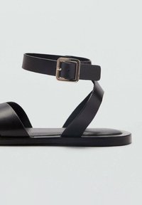 Sandalo in pelle nera con una larga fascia anteriore e una sottile cinghia per la caviglia dotata di una fibbia metallica quadrata. Suola piatta, design minimale.