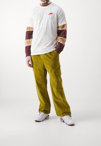 Vit Nike-t-shirt med logga, lager på lager med bruna och beige randiga långärmade tröjor. Olivgröna kordflórdbyxor och vita sneakers med röda accenter.