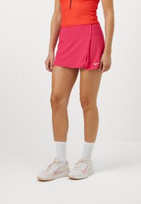 Rózsaszín sport szoknya oldalsó hasítékkal, rugalmas anyagból készült. Fehér zoknikkal és világos rózsaszín Nike cipőkkel viselve, amelyek rózsaszín logóval rendelkeznek.