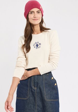 Maglia a maniche lunghe color crema con ricamo di un uccello, abbinata a una gonna di jeans blu con due tasche laterali e dettagli con bottoni. Berretto a coste rosa in testa.