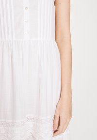 Hvid linned kjole med lodrette plisseringer, en række knapper og blondekant langs kanten. Stoffet ser let og tekstureret ud.