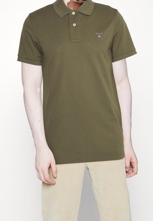 Oliwkowa koszulka polo z guzikami przy kołnierzyku, krótkimi rękawami, miękkim materiałem i małym logo na piersi. Połączona z jasnymi spodniami sztruksowymi.