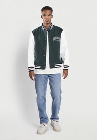 Chaqueta de varsity verde y blanca con rayas negras, cierre de botón y estampado gráfico. Se lleva sobre una camiseta blanca y se combina con jeans azules.