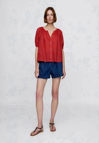 Blusa rossa a fantasia con maniche a sbuffo e bottoni davanti, abbinata a pantaloni corti di jeans. Indossata con sandali piatti.