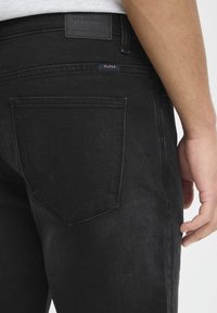 Jeans en denim noir avec une coupe droite. Poche arrière visible et étiquette de marque. Le tissu a une surface lisse avec une légère texture.