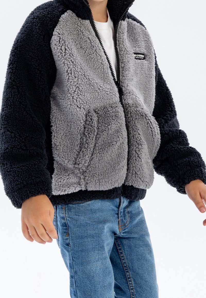 Enfant portant une veste polaire noire et grise zippée avec poches avant et un jean bleu, debout sur fond blanc.