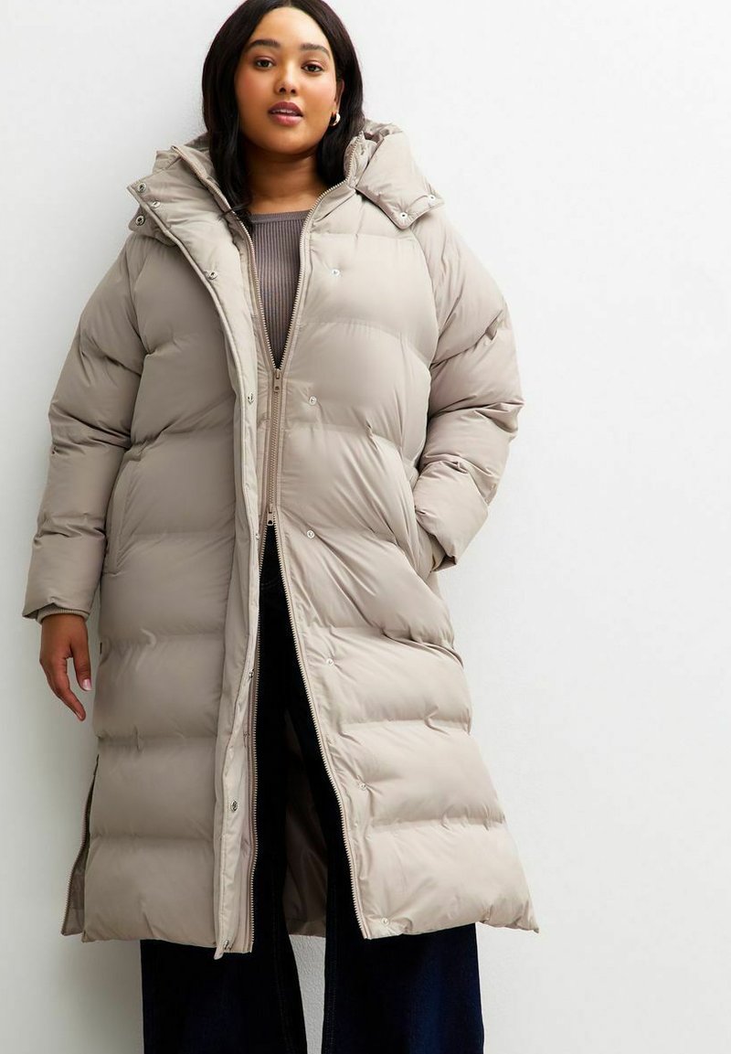 Mujer joven con un abrigo de invierno acolchado largo color beige sobre un suéter marrón y pantalones oscuros, de pie frente a un fondo blanco.
