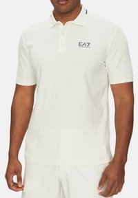 Camisa polo de color claro hecha de algodón, con cuello clásico, tapeta de tres botones y el logo de EA7 en el pecho.