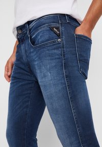 Vaqueros de mezclilla azul oscuro con corte slim, que cuentan con un bolsillo frontal, una etiqueta de logo negra distintiva y costuras en contraste a lo largo de los lados.