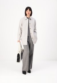 Giacca di lana grigia chiara con chiusura a bottoni sopra un maglione grigio abbinato e pantaloni a gamba larga. La modella tiene in mano una borsa a tracolla nera e crema con texture.