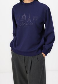 Sweat-shirt bleu marine avec des monuments parisiens imprimés en tons sombres. Encolure ronde, poignets côtelés et coupe décontractée. Pantalon gris visible.