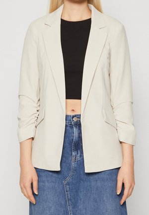 Mujer con blazer beige sobre una camiseta corta negra y falda vaquera azul sobre un fondo liso.