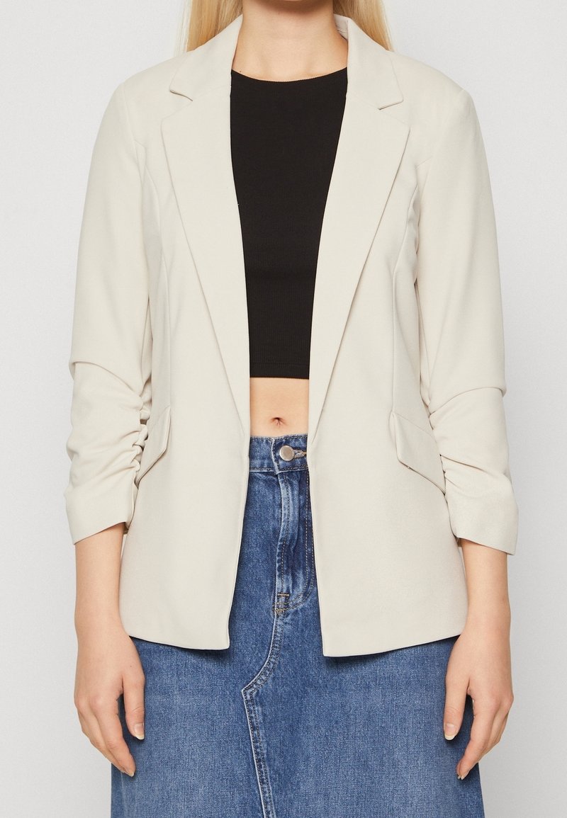 Femme portant un blazer beige sur un crop top noir et une jupe en denim bleue sur un fond uni.