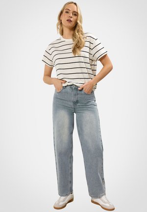 Kobieta ubrana w krótki rękaw w białe i czarne paski, jasnoniebieskie szerokie spodnie dżinsowe i białe trampki, stojąca z rękami w kieszeniach.