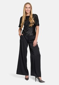 Schwarzes Paillettentop mit kurzen Ärmeln, kombiniert mit weiten schwarzen Hosen. Das Modell trägt metallische High Heels. Glatte Stofftextur.