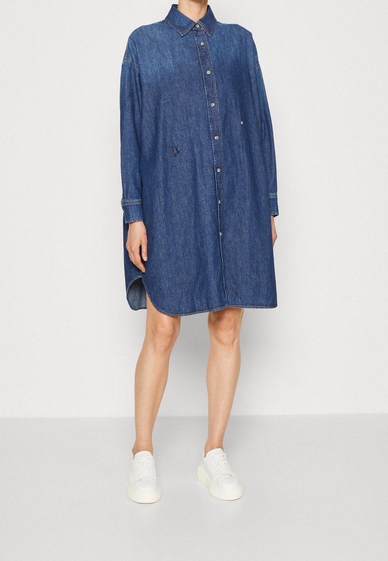 Denim overhemdjurk met een kraag, lange mouwen en een knoopsluiting aan de voorkant. Heeft een afgeronde zoom en een losse pasvorm, gecombineerd met witte sneakers.