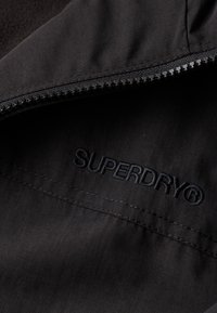 Czarna tkanina o gładkiej fakturze, z zamkiem błyskawicznym i wytłoczonym logo "SUPERDRY" w ciemnym wątku.