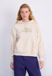 Beige hettegenser med frontlomme og justerbar hette, med en grønn grafikk "new balance" logo. Brukt med lilla bukser.