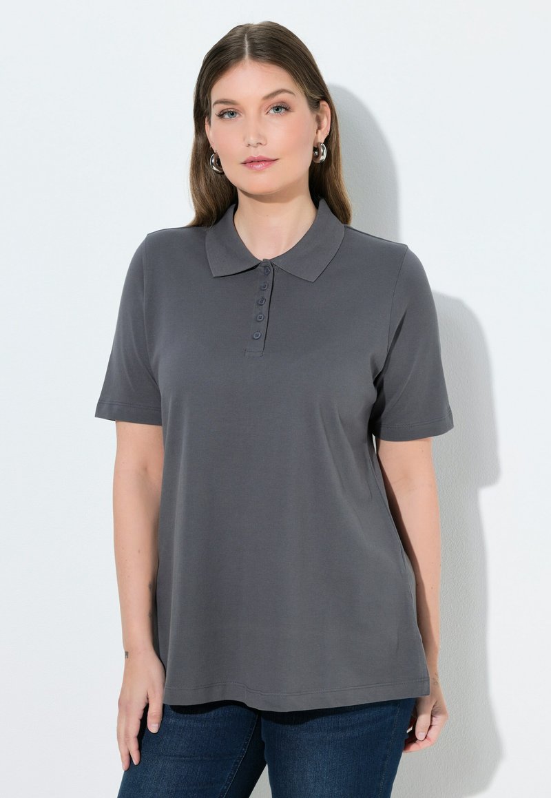 Mujer con cabello largo y castaño, que lleva pendientes de aro plateados, una camiseta polo gris oscuro de manga corta y vaqueros azules, sobre un fondo blanco liso.