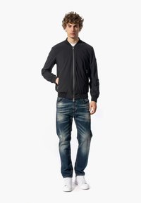 Giacca bomber nera con chiusura a zip e polsini a coste, indossata sopra jeans blu strappati e sneakers bianche.