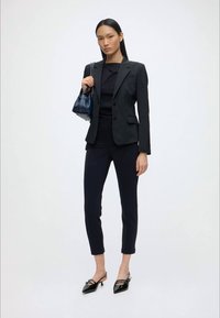 Woman wearing a black tailored blazer, black pants, black top, carrying a navy handbag, and black pointed-toe slingback heels, standing on white background.