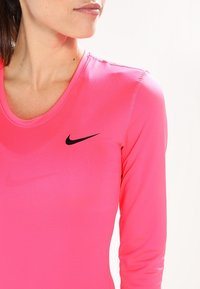 Pink long-sleeve athletic top made of stretchy fabric, featuring a rounded neckline and black Nike logo on the left chest. Subtle stitching details.