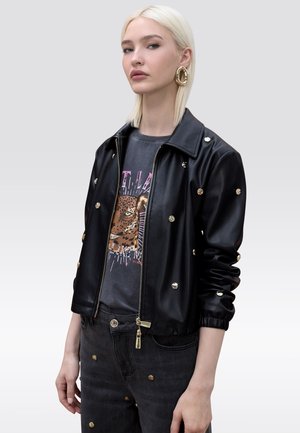 Mujer rubia con chaqueta de cuero negra tachonada, camiseta gráfica con estampado de leopardo, jeans negros tachonados a juego y grandes pendientes de aro dorados.