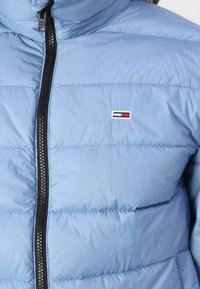 Ljust blå pufferjacka med hög krage, med en svart dragkedja och en liten broderad logga i röd, vit och blå på bröstet.
