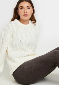 Pull en maille torsadée blanc épais avec un col rond côtelé, associé à des leggings côtelés marron foncé. Textures douces et coupe décontractée.