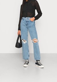Lysblå højtaljede ripped jeans med frynser, parret med en gennemsigtig sort skjorte med knapper og sorte høj-top sneakers, mens der holdes en vævet sort taske.