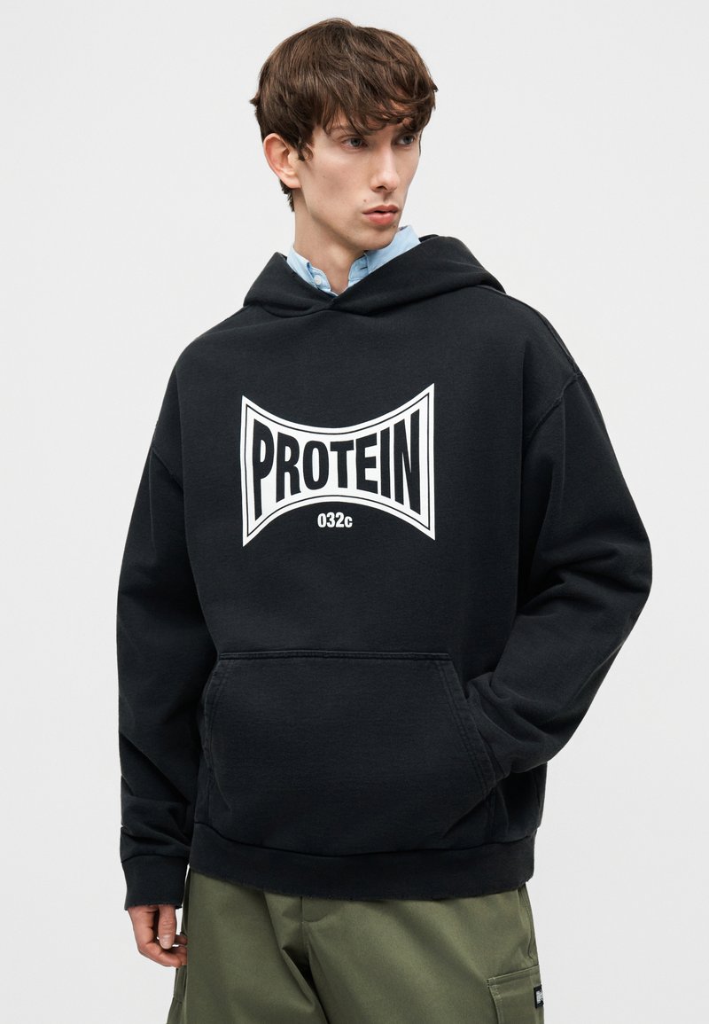Jonge man draagt een zwart capuchontrui met witte tekst "PROTEIN 032c", groene broek, kijkt opzij tegen een effen achtergrond.