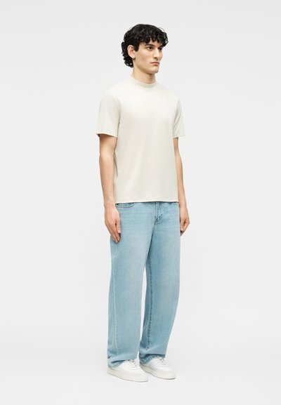 Νέος άνδρας με κατσαρά μαλλιά στέκεται φορώντας απλό μπεζ κοντομάνικο πουκάμισο, φαρδιά τζιν παντελόνια ανοιχτού μπλε χρώματος και λευκά αθλητικά παπούτσια σε λευκό φόντο.