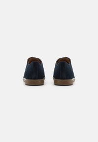 Pantofi slip-on din suede albastru închis, cu vârf rotund, având o talpă din cauciuc maronie deschisă și cusături minime de-a lungul șonului.