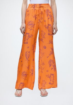 Pantalon orange ample avec motifs floraux violets et visages abstraits, taille élastique avec cordon, porté avec des sandales blanches à lanière.