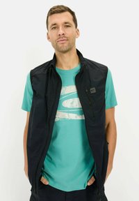 Sort ærmeløs vest med lynlås foran, båret over en turkis t-shirt med et hvidt grafisk print. Glat, letvægtsstof.