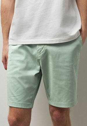 Mand iført lysegrønne shorts og en hvid t-shirt med hænderne i lommerne, vist fra taljen til knæene.