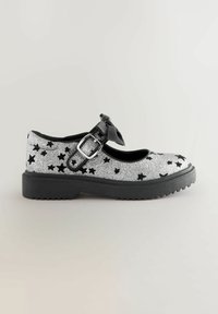 Chaussure Mary Jane en paillettes argentées avec des motifs d'étoiles noires, bout rond, bride de cheville avec un détail en nœud, et semelle en caoutchouc noir.