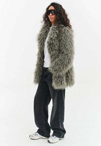 Osoba nosząca oversize'owy szary, puszysty futrzany płaszcz, duże czarne okulary przeciwsłoneczne, białą koszulę, luźne czarne spodnie i białe trampki, stojąca na białym tle.