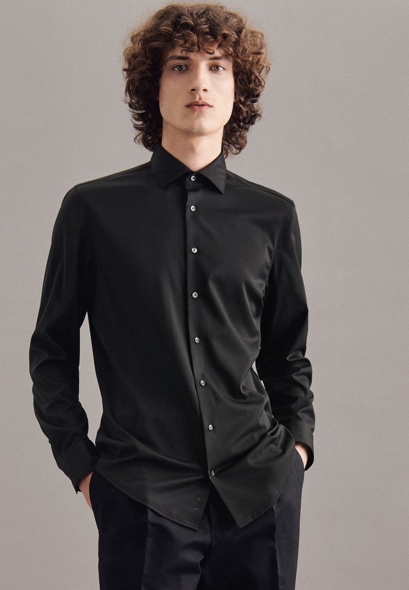 Zwarte longsleeve shirt van gladde stof, met een puntige kraag en subtiele knopen. Het shirt is in donkere broeken gestoken.
