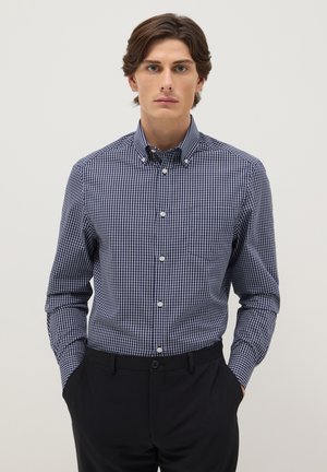 Uomo che indossa una camicia a quadri blu navy e bianca con bottoni e pantaloni neri, in piedi con le mani in tasca davanti a uno sfondo semplice.