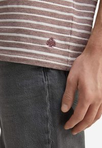 Hnědo-bílá pruhovaná košile s krátkým rukávem a detailní výšivkou stromu na lemu, spárovaná s tmavě šedými džínami.
