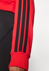 Rode sweatshirt sleeve met zwarte drievoudige streepdetail. De stof lijkt glad en elastisch, met een geribbelde manchette voor een strakke pasvorm.