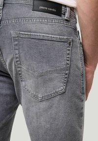 Grijze denim jeans met een close-up weergave van de achterzak, voorzien van een driehoekige flap en een leren label op de tailleband.