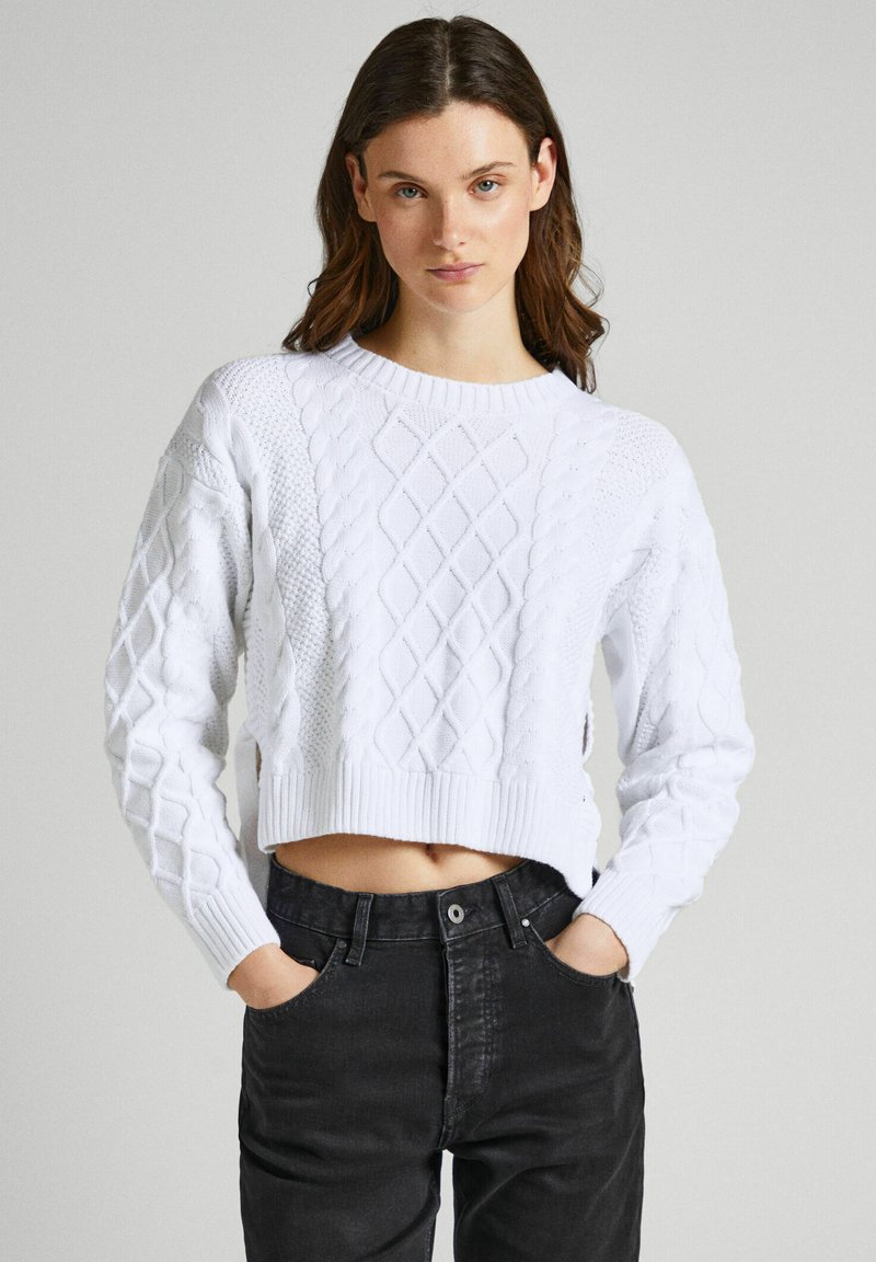 Maglione bianco corto con design a trecce e orlo a coste. Abbinato a jeans neri a vita alta, che mostrano la pancia. Tessuto liscio e strutturato.