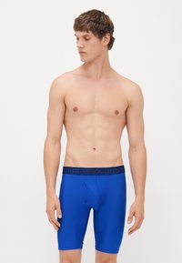 Giovane uomo con i capelli corti che indossa slip lunghi blu Under Armour, in piedi contro uno sfondo chiaro e semplice, che guarda a sinistra.