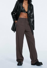 Blazer de cuero negro sobre un crop top negro, combinado con pantalones marrones de pierna ancha y zapatos negros de plataforma gruesa. Texturas suaves, ajuste a medida.