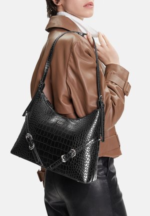 Žena v hnědé kožené bundě a černých kalhotách drží černou texturovanou koženou kabelku s stříbrnými přezkami.